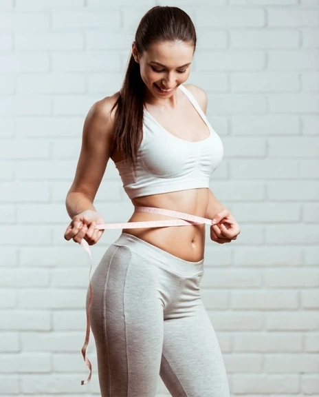 A woman in white athletic wear measuring her waist against a bright white brick wall.